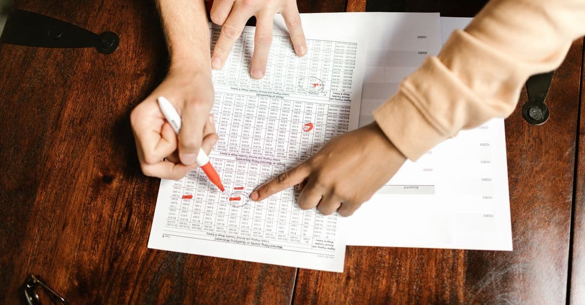 Zwei Hände prüfen Finanzdokumente und markieren Notizen mit rotem Stift auf Holztisch.