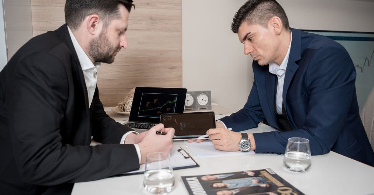 Two businessmen in suits discussing cryptocurrency trends using digital devices in an office setting.