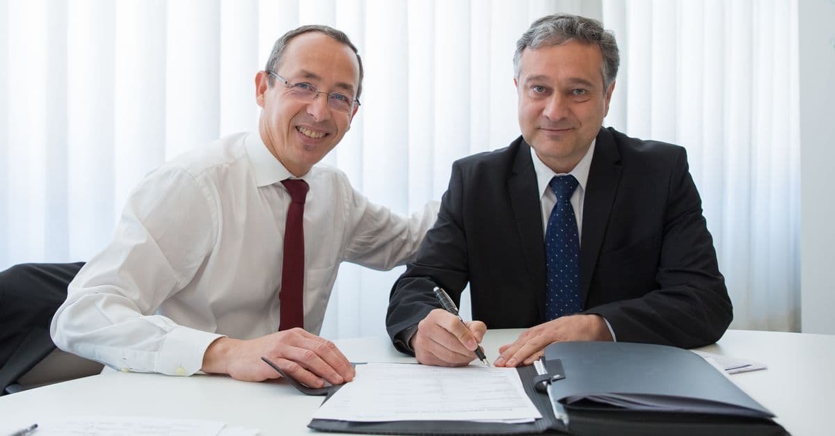 Two businessmen in suits signing a contract at a well-lit office table.