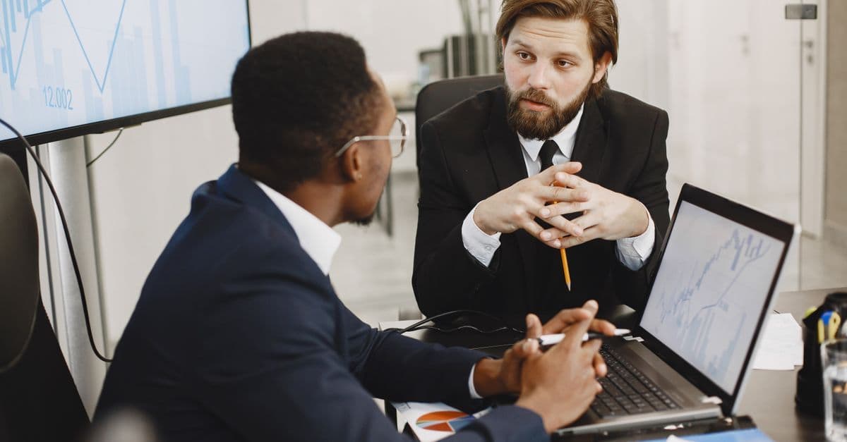 Zwei Geschäftsleute im Anzug besprechen Finanzdaten an Laptops in einem modernen Büro.
