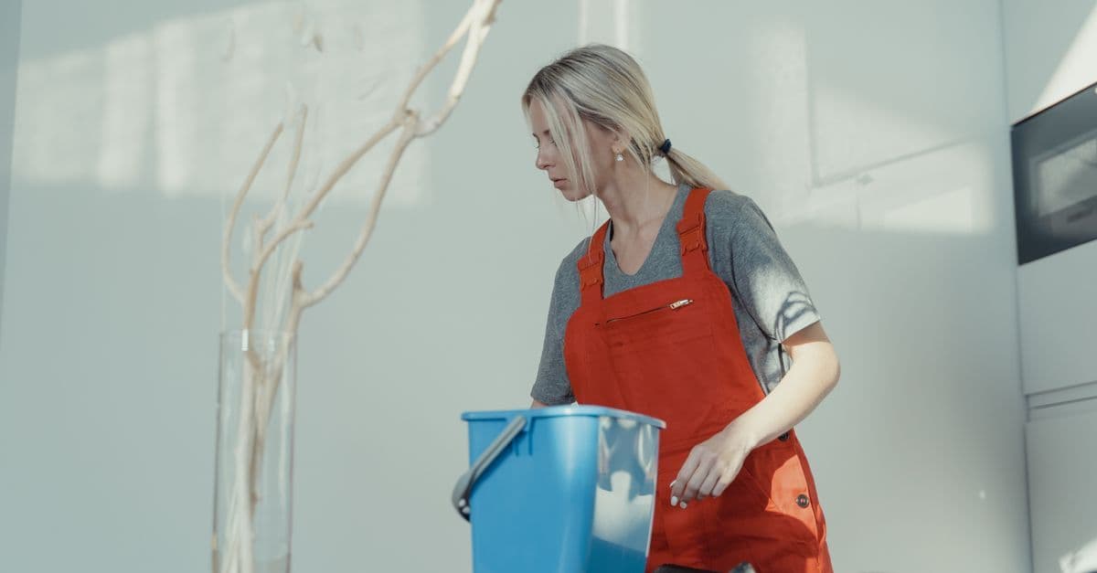 A woman in a red uniform cleans indoors with a blue bucket, ensuring a spotless environment.