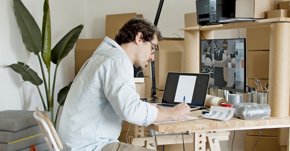 Mann im Homeoffice arbeitet am Laptop, umgeben von Kartons und Verpackungsmaterial für seinen Onlineshop.
