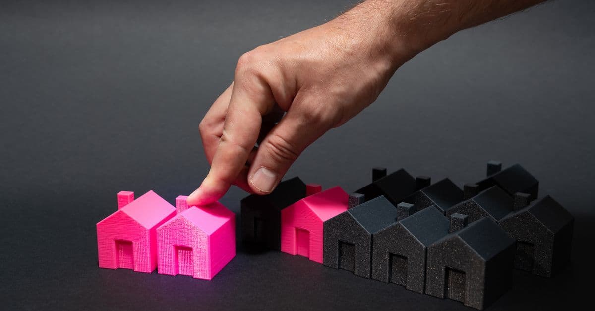 A hand choosing a vibrant pink house model among black miniatures, symbolizing real estate selection.