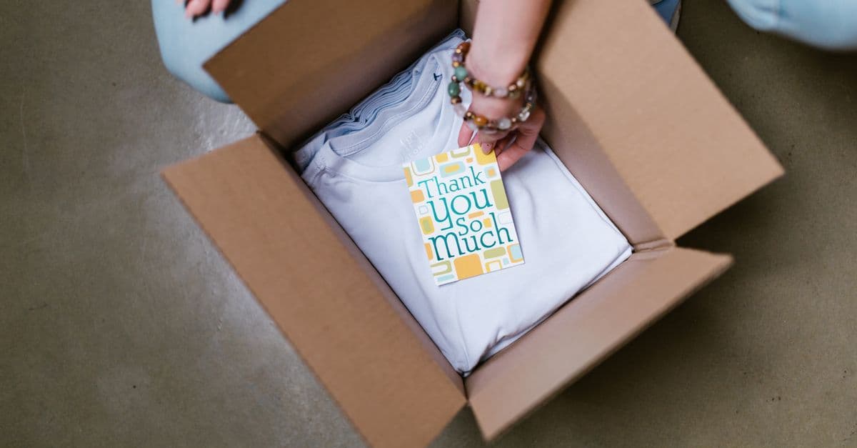 Person legt weißes T-Shirt mit Dankeskarte in einen Karton.