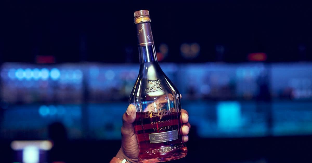 A close-up of a hand holding a cognac bottle against a vibrant bar backdrop.