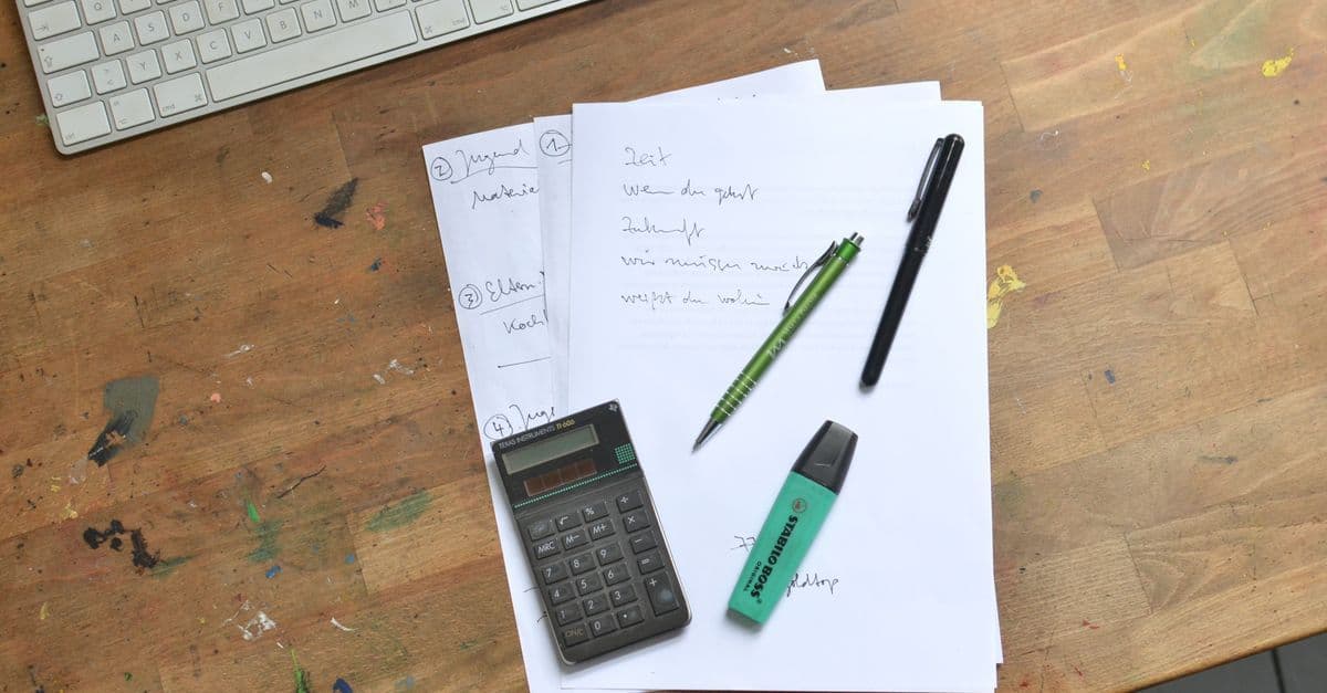 Top view of paper sheets with notes placed with pens with calculator and marker near keyboard on dirty table