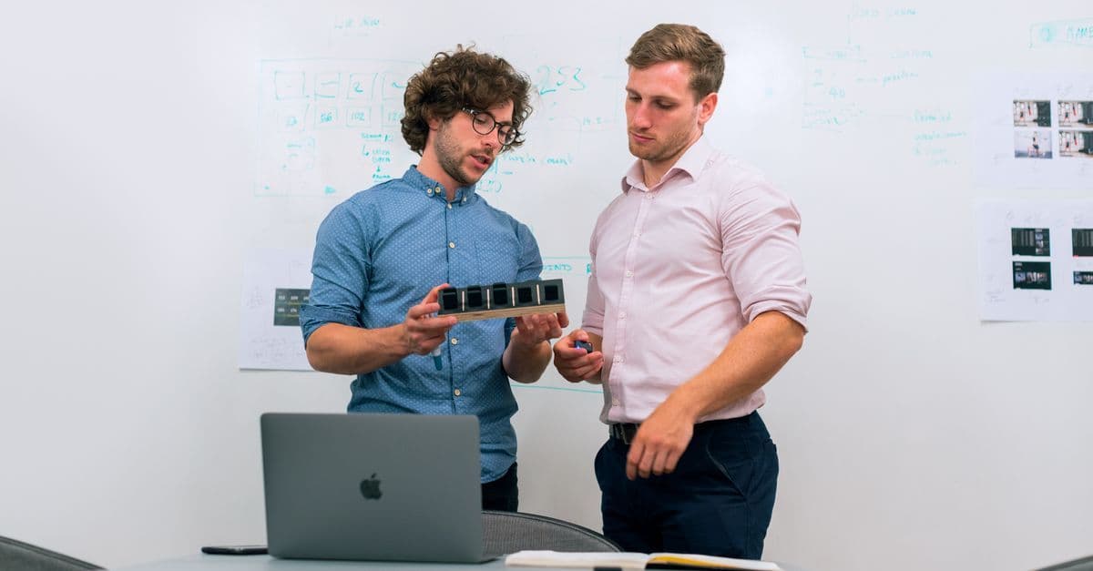 Zwei Männer besprechen im Büro einen technischen Prototypen und prüfen Unterlagen.